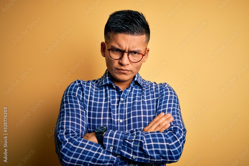 Young handsome latin man wearing casual shirt and glasses over yellow background skeptic and nervous, disapproving expression on face with crossed arms. Negative person.