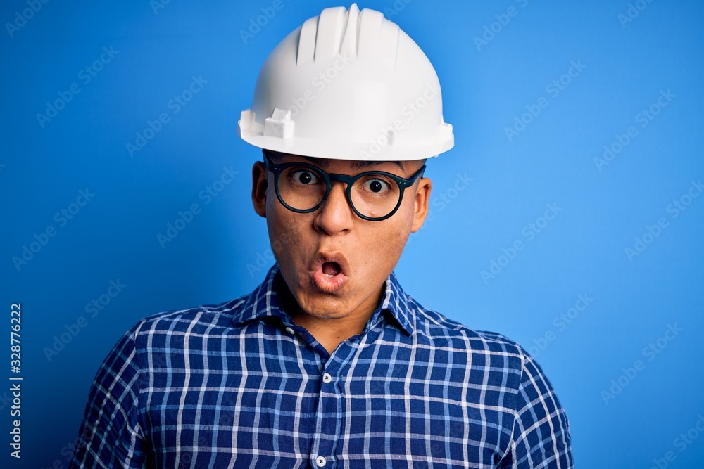 Young handsome engineer latin man wearing safety helmet over isolated blue background afraid and shocked with surprise expression, fear and excited face.