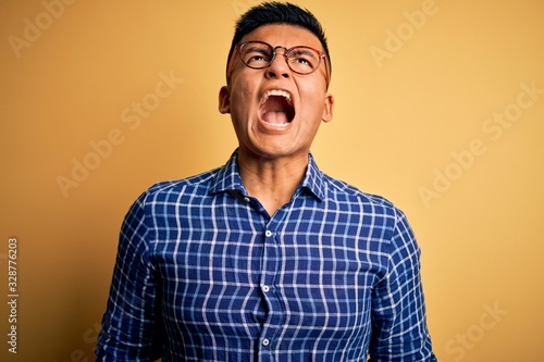 Young handsome latin man wearing casual shirt and glasses over yellow background angry and mad screaming frustrated and furious, shouting with anger. Rage and aggressive concept.