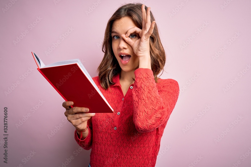 Young blonde student girl reading a book over pink isolated background with happy face smiling doing ok sign with hand on eye looking through fingers Young blonde student girl reading a book over pink isolated background with happy face smiling doing ok sign with hand on eye looking through fingers