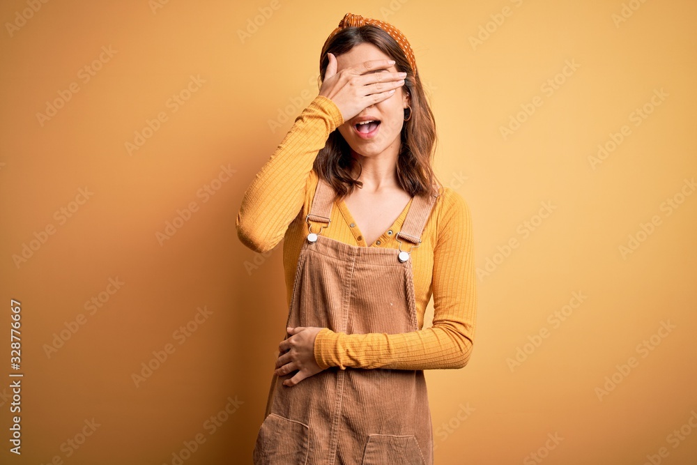 Young beautiful blonde girl wearing overall standing over yellow isolated background smiling and laughing with hand on face covering eyes for surprise. Blind concept. Young beautiful blonde girl wearing overall standing over yellow isolated background smiling and laughing with hand on face covering eyes for surprise. Blind concept.