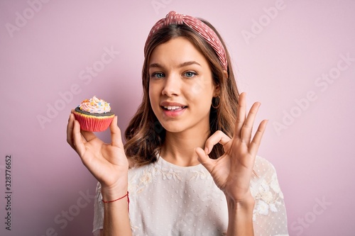 Young blonde girl eating a sweet chocolate cupcake over pink isolated background doing ok sign with fingers  excellent symbol