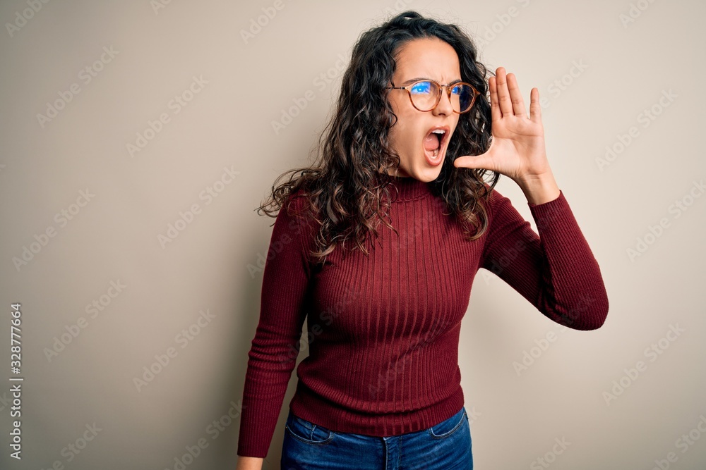 Beautiful woman with curly hair wearing casual sweater and glasses over white background shouting and screaming loud to side with hand on mouth. Communication concept.