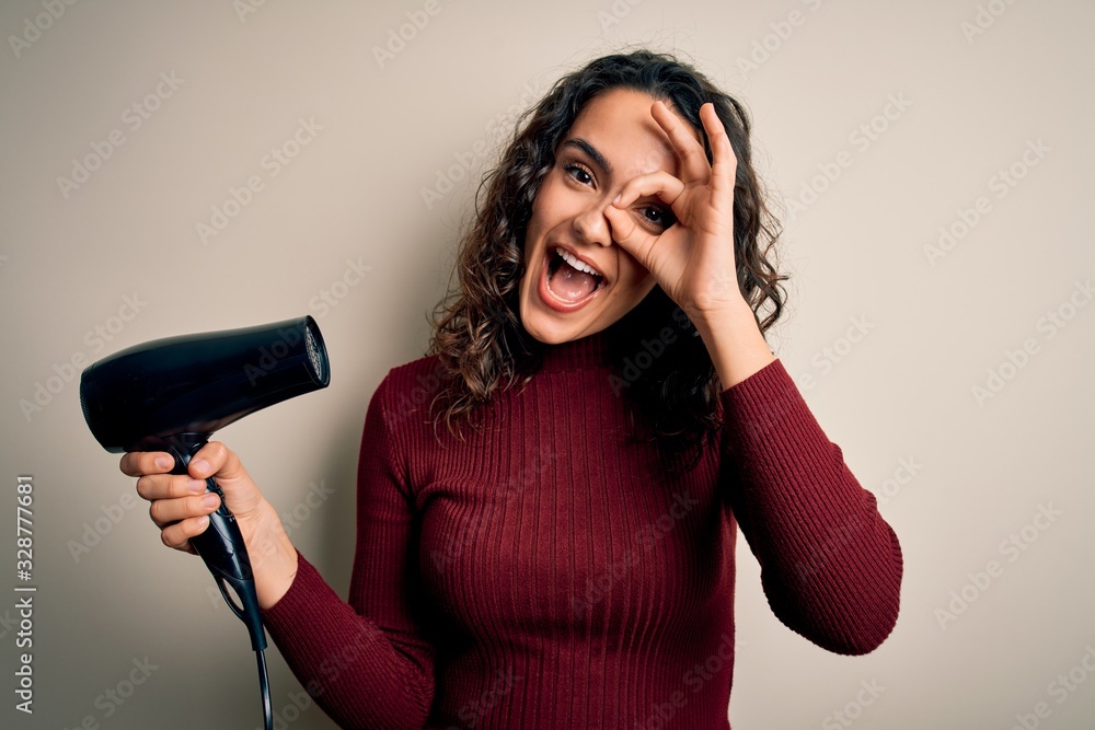 Young beautiful woman with curly hair using hair dryer over isolated white background with happy face smiling doing ok sign with hand on eye looking through fingers Young beautiful woman with curly hair using hair dryer over isolated white background with happy face smiling doing ok sign with hand on eye looking through fingers