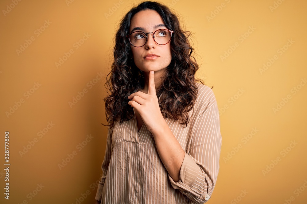 Beautiful woman with curly hair wearing striped shirt and glasses over yellow background Thinking concentrated about doubt with finger on chin and looking up wondering Beautiful woman with curly hair wearing striped shirt and glasses over yellow background Thinking concentrated about doubt with finger on chin and looking up wondering