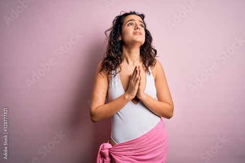Beautiful woman with curly hair on vacation wearing white swimsuit over pink background begging and praying with hands together with hope expression on face very emotional and worried. Begging. © Krakenimages.com