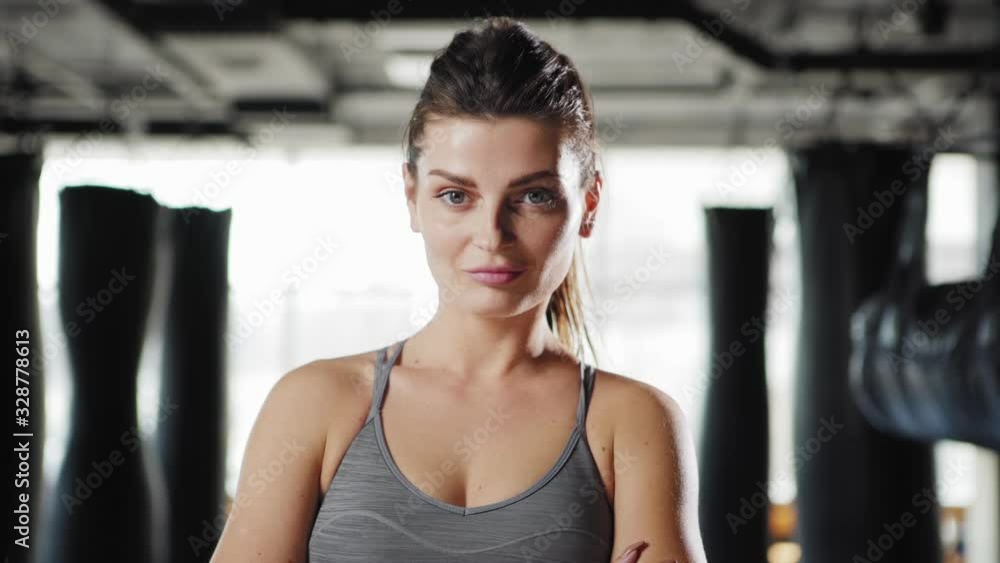 Fade in shot of attractive fit young woman walking towards camera and posing cross armed looking at camera confidently in boxing gym with heavy bags in background Fade in shot of attractive fit young woman walking towards camera and posing cross armed looking at camera confidently in boxing gym with heavy bags in background
