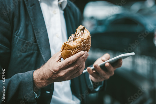Fotografija Man in a blue sute working online on his smartphone while lunchbreak eating a hamburger or sandwich standing outside of business center