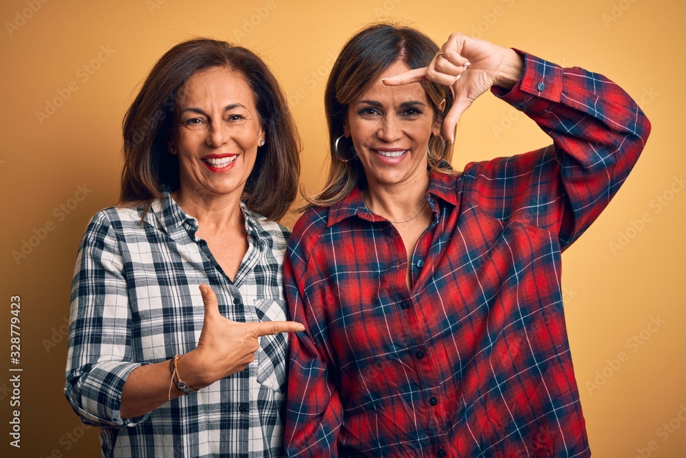 Middle age beautiful couple of sisters wearing casual shirt over isolated yellow background smiling making frame with hands and fingers with happy face. Creativity and photography concept. Middle age beautiful couple of sisters wearing casual shirt over isolated yellow background smiling making frame with hands and fingers with happy face. Creativity and photography concept.