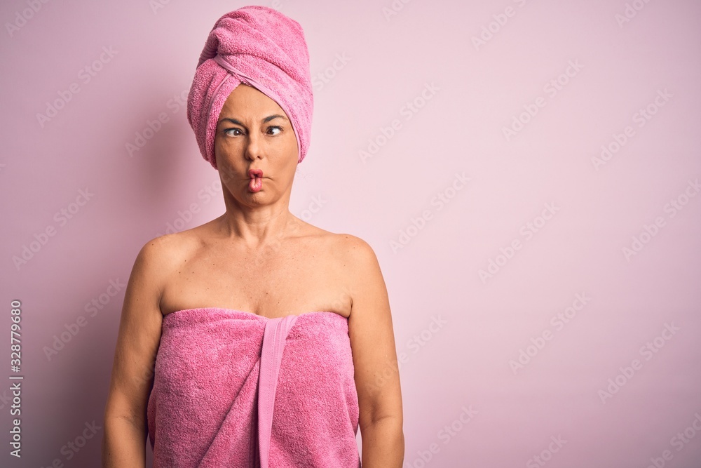 Middle age woman wearing bath towel from beauty body care over pink background making fish face with lips, crazy and comical gesture. Funny expression. Middle age woman wearing bath towel from beauty body care over pink background making fish face with lips, crazy and comical gesture. Funny expression.