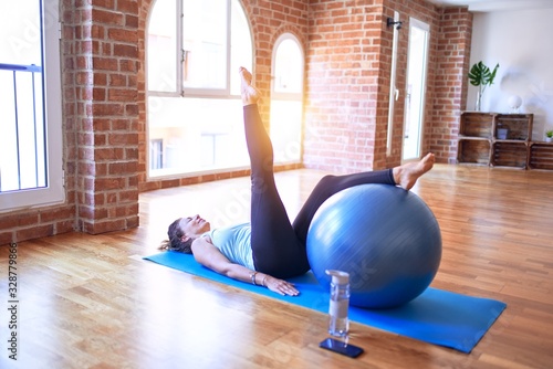 Middle age beautiful sportswoman smiling happy. Doing exercise using fitness ball in a class of yoga at gym