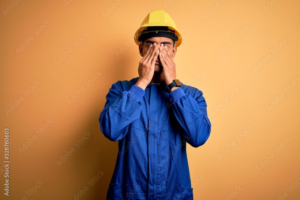 Young handsome african american worker man wearing blue uniform and security helmet rubbing eyes for fatigue and headache, sleepy and tired expression. Vision problem Young handsome african american worker man wearing blue uniform and security helmet rubbing eyes for fatigue and headache, sleepy and tired expression. Vision problem