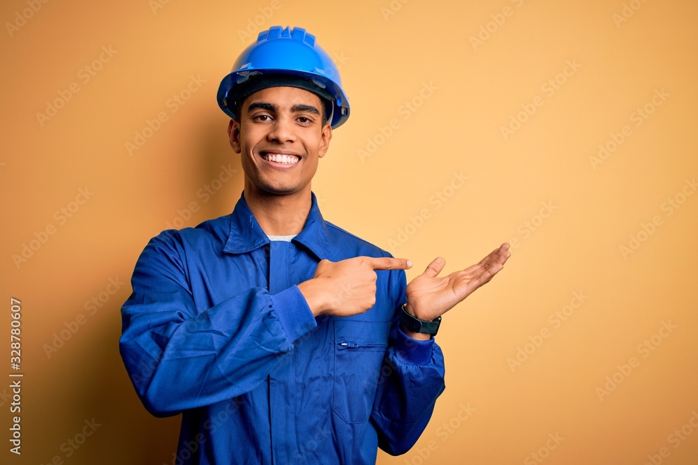 Young handsome african american worker man wearing blue uniform and security helmet amazed and smiling to the camera while presenting with hand and pointing with finger.