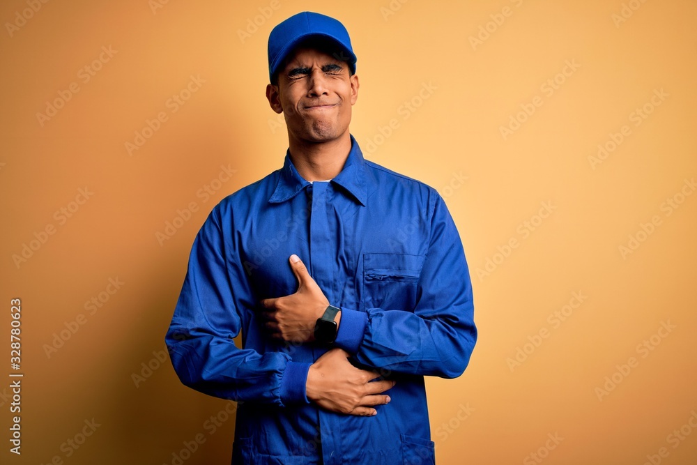 Young african american mechanic man wearing blue uniform and cap over yellow background with hand on stomach because indigestion, painful illness feeling unwell. Ache concept.