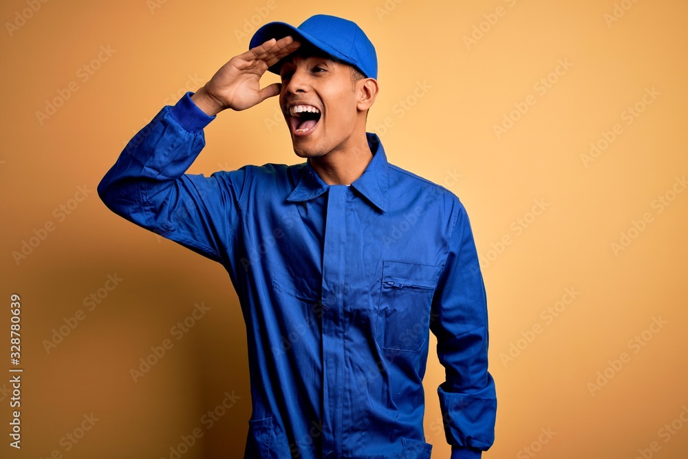 Young african american mechanic man wearing blue uniform and cap over yellow background very happy and smiling looking far away with hand over head. Searching concept.
