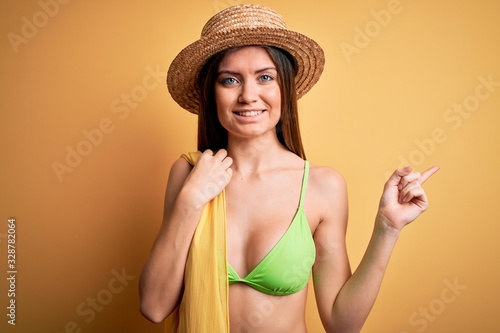 Young beautiful woman with blue eyes on vacation wearing bikini and hat holding towel very happy pointing with hand and finger to the side