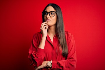 Young beautiful brunette woman wearing casual shirt and glasses over red background with hand on chin thinking about question, pensive expression. Smiling with thoughtful face. Doubt concept.