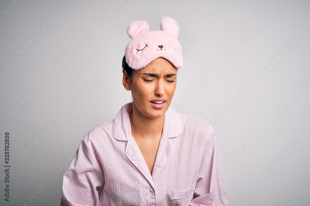 Young beautiful brunette woman wearing pajama and sleep mask over white background with hand on stomach because nausea, painful disease feeling unwell. Ache concept. Young beautiful brunette woman wearing pajama and sleep mask over white background with hand on stomach because nausea, painful disease feeling unwell. Ache concept.