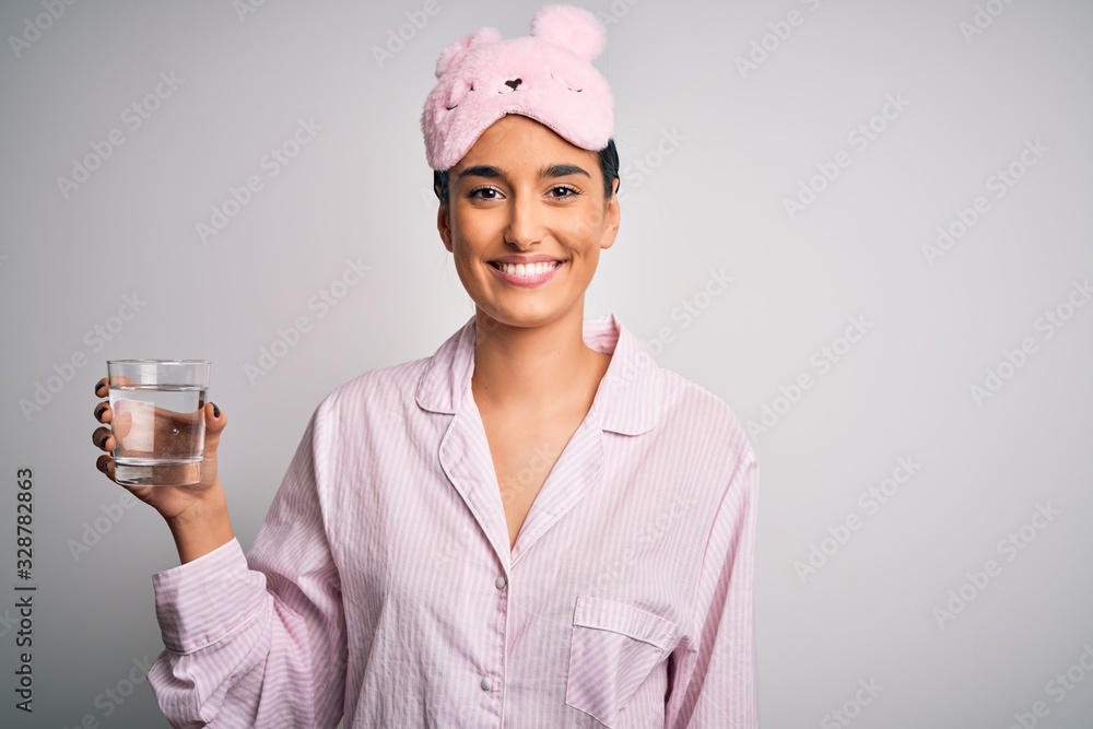 Young beautiful woman wearing pajama and sleep mask drinking glass of water with a happy face standing and smiling with a confident smile showing teeth Young beautiful woman wearing pajama and sleep mask drinking glass of water with a happy face standing and smiling with a confident smile showing teeth
