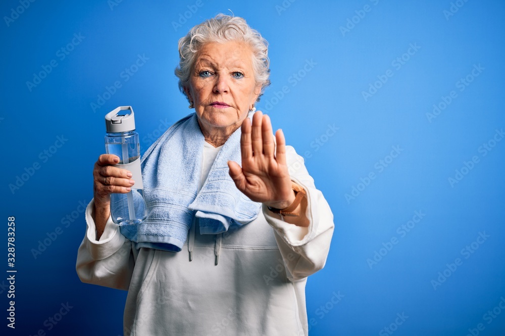 Senior beautiful sporty woman holding bottle of water standing over isolated blue background with open hand doing stop sign with serious and confident expression, defense gesture Senior beautiful sporty woman holding bottle of water standing over isolated blue background with open hand doing stop sign with serious and confident expression, defense gesture