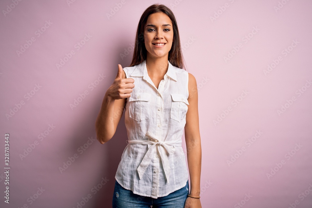 Young beautiful brunette woman wearing casual shirt standing over pink background doing happy thumbs up gesture with hand. Approving expression looking at the camera showing success.