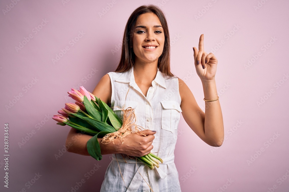 Young beautiful brunette woman holding bouquet of tulips flowers over pink background smiling amazed and surprised and pointing up with fingers and raised arms. Young beautiful brunette woman holding bouquet of tulips flowers over pink background smiling amazed and surprised and pointing up with fingers and raised arms.