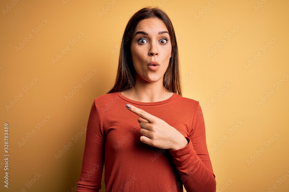Obraz premium Young beautiful brunette woman wearing casual t-shirt standing over yellow background Surprised pointing with finger to the side, open mouth amazed expression.