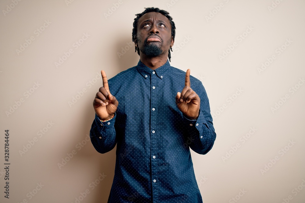 Plakat Young handsome african american man wearing casual shirt standing over white background Pointing up looking sad and upset, indicating direction with fingers, unhappy and depressed.
