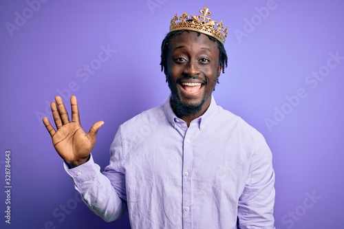 Young african american man wearing golden crown of king over isolated purple background showing and pointing up with fingers number five while smiling confident and happy.