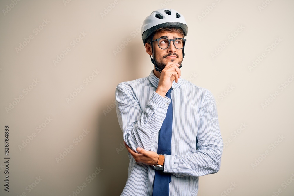 Young businessman wearing glasses and bike helmet standing over isolated white bakground with hand on chin thinking about question, pensive expression. Smiling with thoughtful face. Doubt concept. Young businessman wearing glasses and bike helmet standing over isolated white bakground with hand on chin thinking about question, pensive expression. Smiling with thoughtful face. Doubt concept.
