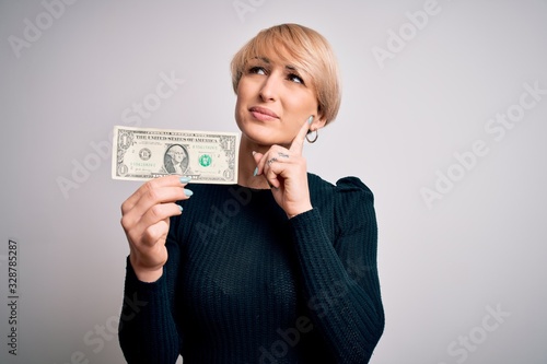 Young blonde woman with short hair holding one dollar banknote over isolated background serious face thinking about question, very confused idea
