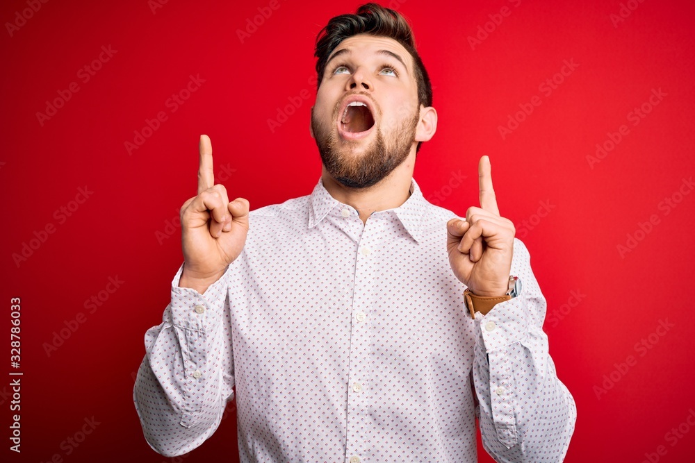 Young blond businessman with beard and blue eyes wearing elegant shirt over red background amazed and surprised looking up and pointing with fingers and raised arms. Young blond businessman with beard and blue eyes wearing elegant shirt over red background amazed and surprised looking up and pointing with fingers and raised arms.