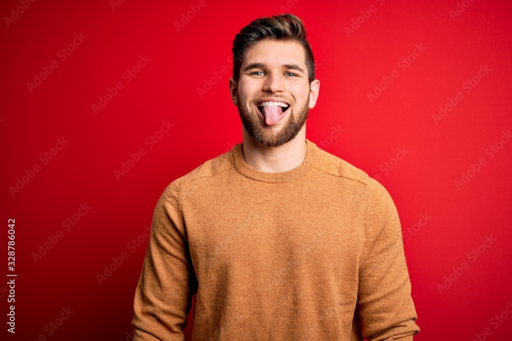 Young blond man with beard and blue eyes wearing casual s over red background sticking tongue out happy with funny expression. Emotion concept. Young blond man with beard and blue eyes wearing casual s over red background sticking tongue out happy with funny expression. Emotion concept.