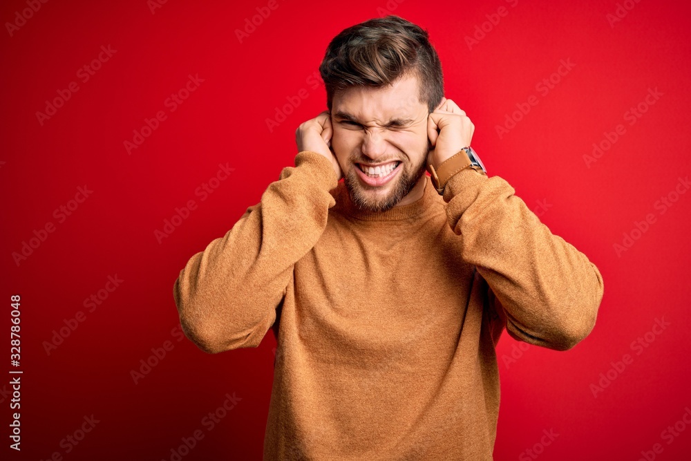 Young blond man with beard and blue eyes wearing casual sweater over red background covering ears with fingers with annoyed expression for the noise of loud music. Deaf concept. Young blond man with beard and blue eyes wearing casual sweater over red background covering ears with fingers with annoyed expression for the noise of loud music. Deaf concept.