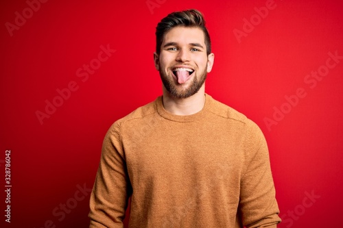 Young blond man with beard and blue eyes wearing casual s over red background sticking tongue out happy with funny expression. Emotion concept.
