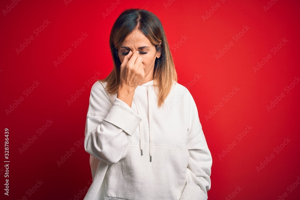 Middle age beautiful sportswoman wearing casual sweatshirt over isolated red background tired rubbing nose and eyes feeling fatigue and headache. Stress and frustration concept.