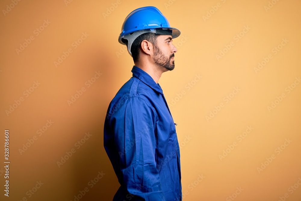 Mechanic man with beard wearing blue uniform and safety helmet over yellow background looking to side, relax profile pose with natural face and confident smile. Mechanic man with beard wearing blue uniform and safety helmet over yellow background looking to side, relax profile pose with natural face and confident smile.