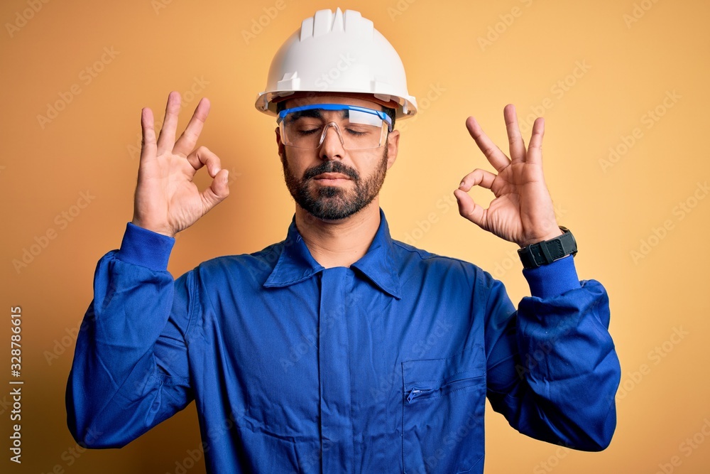Mechanic man with beard wearing blue uniform and safety glasses over yellow background relax and smiling with eyes closed doing meditation gesture with fingers. Yoga concept.