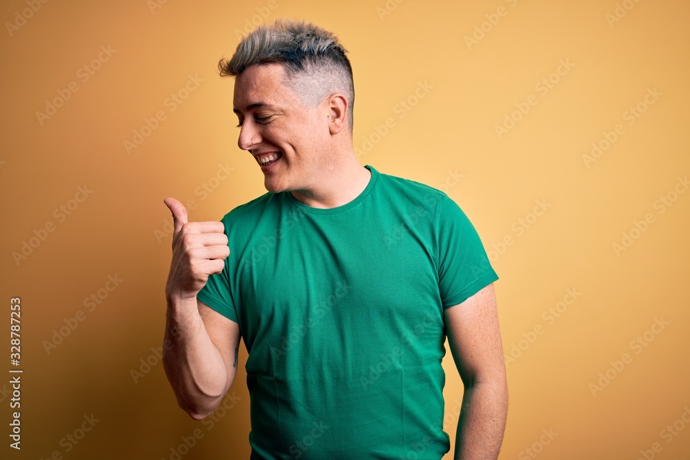 Young handsome modern man wearing casual green t-shirt over yellow background smiling with happy face looking and pointing to the side with thumb up. Young handsome modern man wearing casual green t-shirt over yellow background smiling with happy face looking and pointing to the side with thumb up.