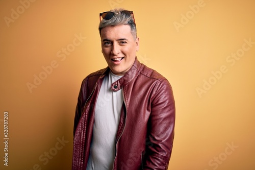 Young handsome modern man wearing fashion leather jacket and sunglasses over yellow background winking looking at the camera with sexy expression, cheerful and happy face.