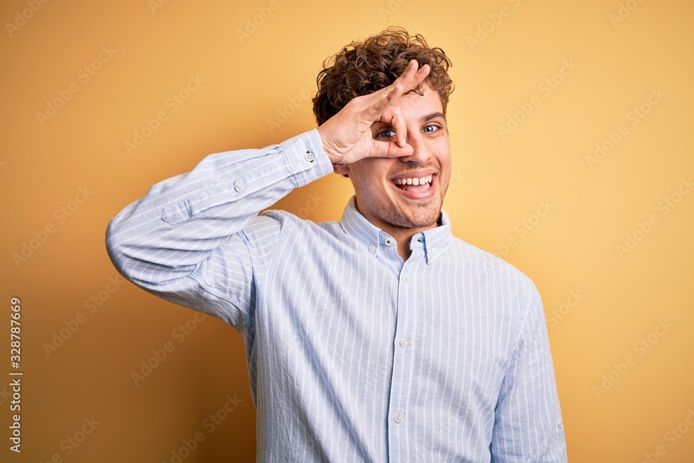 Young blond handsome man with curly hair wearing striped shirt over yellow background doing ok gesture with hand smiling, eye looking through fingers with happy face. Young blond handsome man with curly hair wearing striped shirt over yellow background doing ok gesture with hand smiling, eye looking through fingers with happy face.