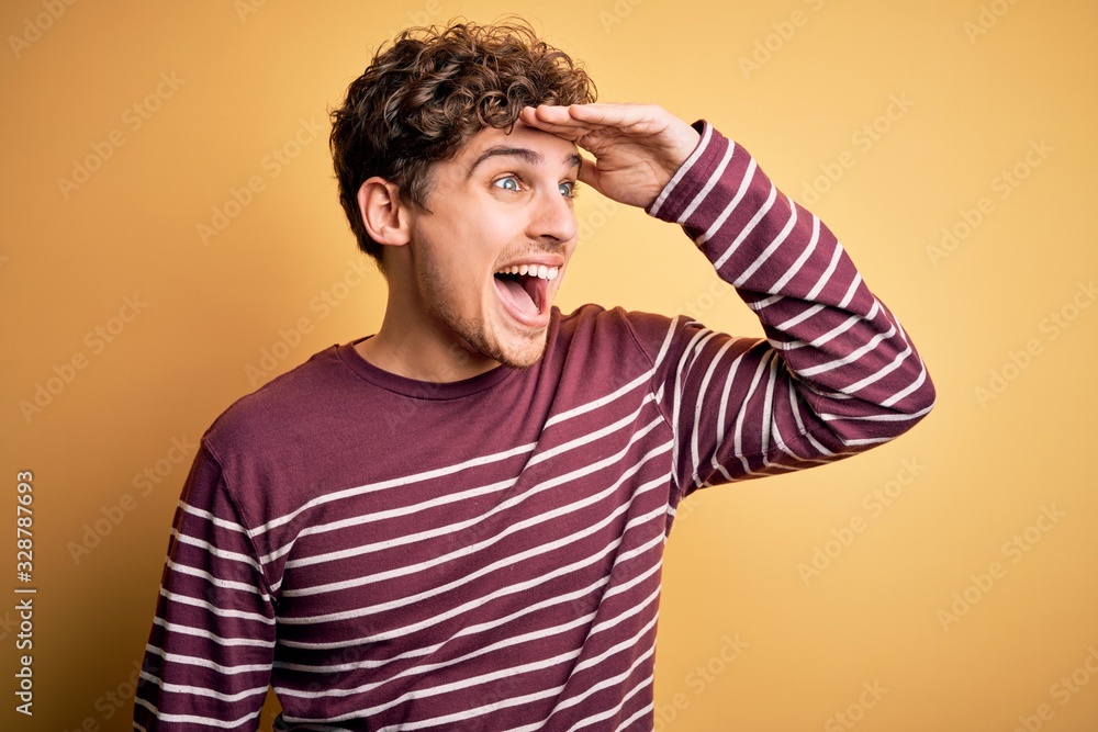 Young blond handsome man with curly hair wearing casual striped sweater very happy and smiling looking far away with hand over head. Searching concept. Young blond handsome man with curly hair wearing casual striped sweater very happy and smiling looking far away with hand over head. Searching concept.