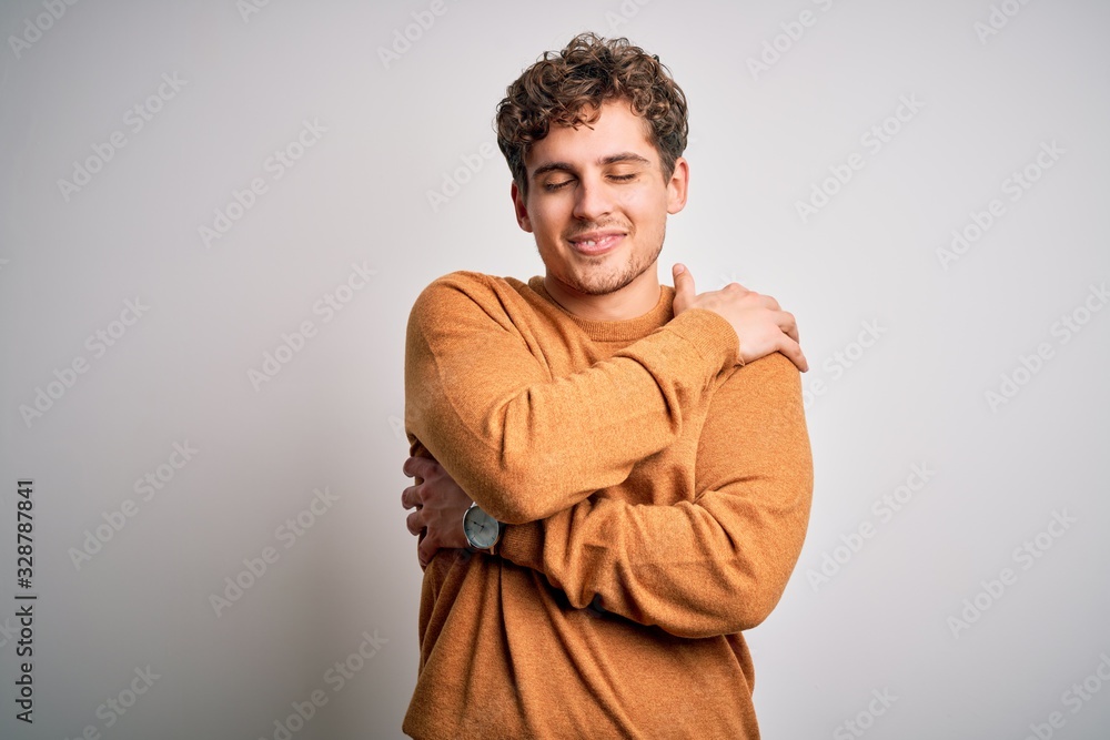 Young blond handsome man with curly hair wearing casual sweater over white background Hugging oneself happy and positive, smiling confident. Self love and self care