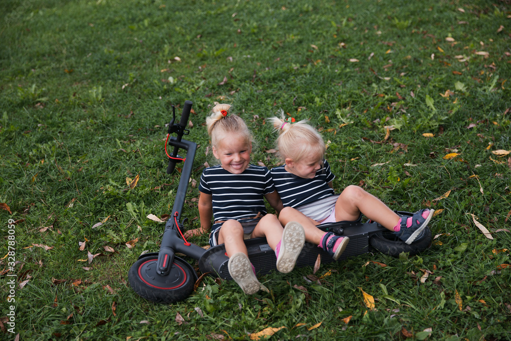 Fototapeta premium Twin blonde little girls sit on fallen electric scooter in green park