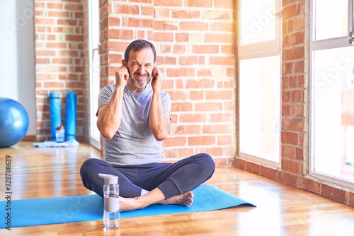 Middle age handsome sportman sitting on mat doing stretching yoga exercise at gym covering ears with fingers with annoyed expression for the noise of loud music. Deaf concept.