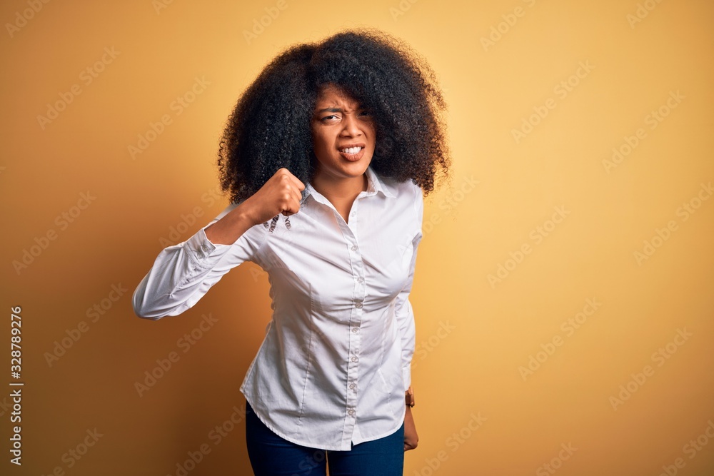 Young beautiful african american elegant woman with afro hair standing over yellow background angry and mad raising fist frustrated and furious while shouting with anger. Rage and aggressive concept. Young beautiful african american elegant woman with afro hair standing over yellow background angry and mad raising fist frustrated and furious while shouting with anger. Rage and aggressive concept.
