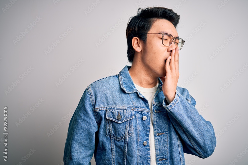 Young handsome chinese man wearing denim jacket and glasses over white background bored yawning tired covering mouth with hand. Restless and sleepiness.