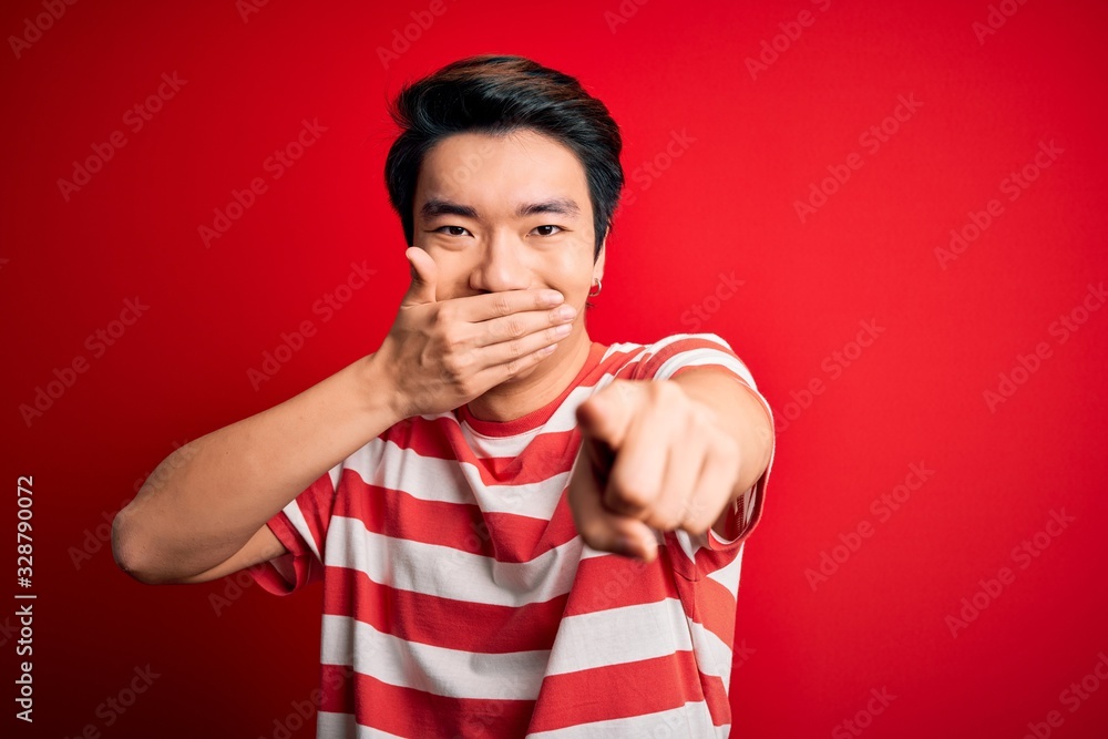 Young handsome chinese man wearing casual striped t-shirt standing over red background laughing at you, pointing finger to the camera with hand over mouth, shame expression Young handsome chinese man wearing casual striped t-shirt standing over red background laughing at you, pointing finger to the camera with hand over mouth, shame expression