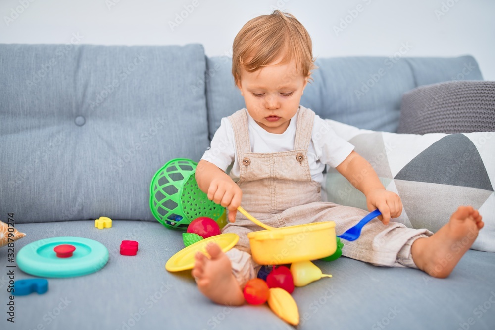 Adorable blonde toddler sitting on the sofa playing with plastic meals toys at home Adorable blonde toddler sitting on the sofa playing with plastic meals toys at home