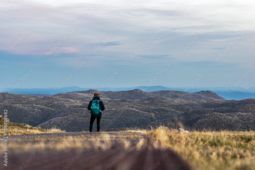 Moody low angle evening view of a young female hiker standing with the mountain landscape near the summit of Mount Kosciuszko, Australia's highest summit in the Snowy Mountains, in the background.